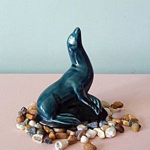 Könnte beinhalten: Eine tealblaue Keramik-Seehundsfigur, die auf einem Bett aus kleinen braunen, weißen und beigen Steinen sitzt.