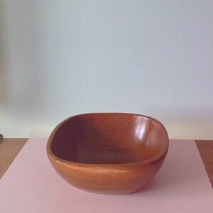 May include: A brown wooden bowl with a rounded square shape. The bowl is sitting on a pink surface.