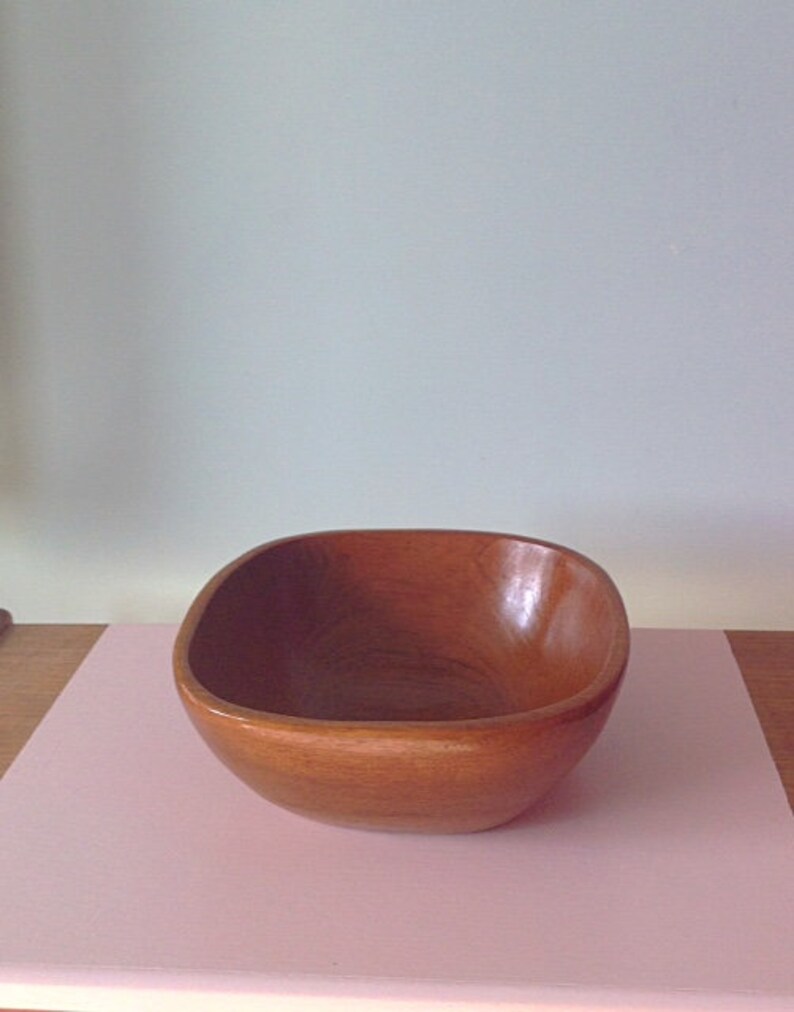 May include: A brown wooden bowl with a rounded square shape. The bowl is sitting on a pink surface.