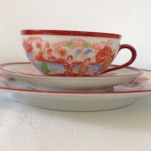 Könnte beinhalten: Ein Vintage-Teeservice, bestehend aus einer Teetasse, Untertasse und Teller. Das Set hat einen roten Rand und eine detaillierte Landschaftsszene mit Figuren in Rot, Blau und Grün. Das Set steht auf einer weißen Oberfläche.