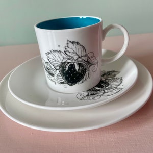 May include: A white teacup and saucer set with a black and white illustration of a strawberry. The teacup has a teal blue interior.