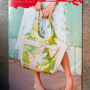 May include: A sewing pattern package for a market bag by Heather Bailey. The cover features a green and white floral tote bag with a bow detail. The text reads Heather Bailey Sewing Patterns, Saturday Market Bag, and a stylish, expandable shopping tote.