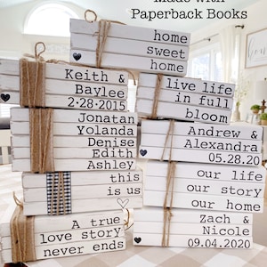 May include: A stack of white painted books with black text, tied with twine, and decorated with small black hearts. The books are stacked to create a decorative display with personalized messages, names, and dates. The books are arranged on a white and brown checkered surface.