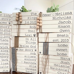 May include: A stack of white paperback books with names and dates written on them. The books are tied together with twine and decorated with wooden beads and a small black and white checkered ribbon. The books are arranged in a tower with the words "Made with Paperback Books" written on the table below.