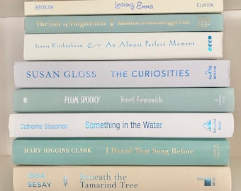 May include: A stack of hardcover books in various shades of blue, white, and beige. Titles and authors are visible, including "Anarchy and Old Dogs" by Colin Cotterill and "Ice and Stone" by Marcia Muller. The books are neatly arranged on a shelf.