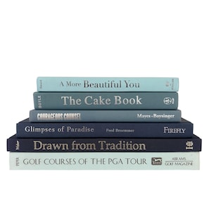 May include: A stack of six hardcover books with spines in shades of blue, ranging from light aqua to deep navy. The titles include A More Beautiful You, The Cake Book, Courageous Counsel, Glimpses of Paradise, Drawn from Tradition, and Golf Courses of the PGA Tour.
