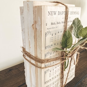 May include: A stack of vintage hymnals tied with twine and a sprig of greenery. The hymnal cover features the text "NEW BAPTIST HYMNAL" and the title of a song "Holy, Holy, Holy, Lord God Almighty".