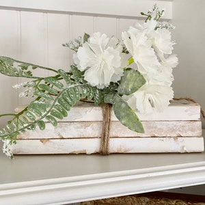 May include: A stack of white books with distressed covers are tied together with twine and decorated with a bouquet of white flowers and green foliage.