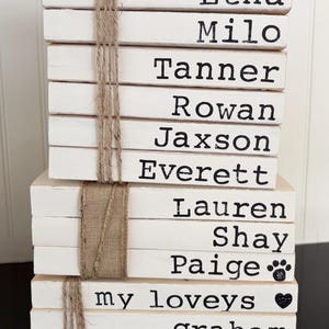 May include: A stack of eleven white rectangular wooden blocks tied with twine and burlap ribbon. Each block features black printed text including the names Etna, Milo, Tanner, Rowan, Jaxson, Everett, Lauren, Shay, Paige with a paw print, my loveys with a heart, graham, and charlotte.