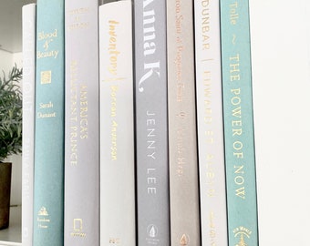 May include: A bookshelf with a collection of books in various shades of blue and gray. The titles of the books include "The Power of Now", "Dunbar", "Anna K", "Inventory", "America's Reluctant Prince", "Blood & Beauty", and "Out Loud".