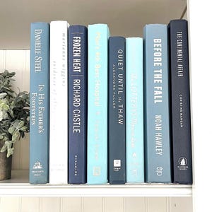 May include: A bookshelf with a collection of blue and white hardcover books. The books are arranged in a row and have titles such as "Frozen Heat", "Quiet Until the Thaw", and "Before the Fall".