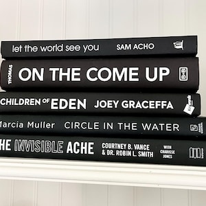 May include: A stack of five hardcover books with black covers and white lettering. Titles include "Let the World See You," "On the Come Up," "Children of Eden," "Circle in the Water," and "The Invisible Ache." The books are arranged on a white shelf.