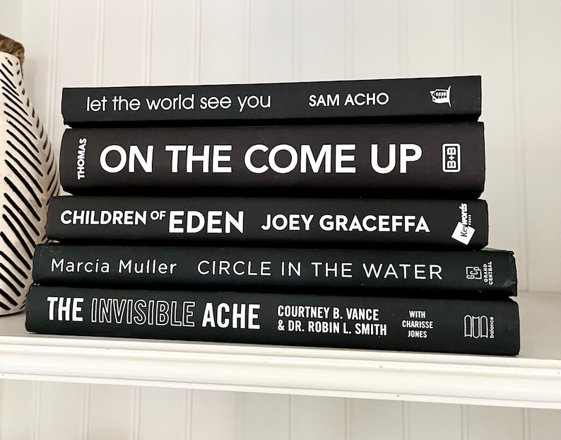 May include: A stack of five hardcover books with black covers and white lettering. Titles include "Let the World See You," "On the Come Up," "Children of Eden," "Circle in the Water," and "The Invisible Ache." The books are arranged on a white shelf.