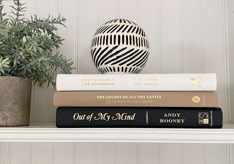 May include: A stack of three books on a white shelf. The top book is white with gold lettering that reads "The Ensemble" and "Ava Garbel". The middle book is beige with gold lettering that reads "The Colors of All the Cattle" and "Alexander McCall Smith". The bottom book is black with gold lettering that reads "Out of My Mind" and "Andy Rooney". A black and white geometric sphere sits on top of the books.