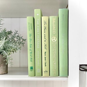 May include: A shelf arrangement featuring a collection of books with light green covers. Titles include "The Star Side of Bird Hill," "The First Mistake," and "The Allure of Order." A potted plant with green foliage sits to the left, adding a touch of nature.