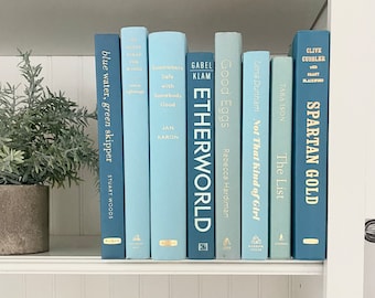 May include: A collection of hardcover books in shades of blue and green, arranged on a white shelf. Titles include "blue water, green skipper", "ETHERWORLD", and "SPARTAN GOLD". A potted plant is visible on the left.