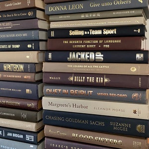 May include: A stack of hardcover books in various colors, including brown, navy, and gray. The titles and authors are printed in gold lettering. The books are arranged in a neat pile, showcasing a collection of literature.