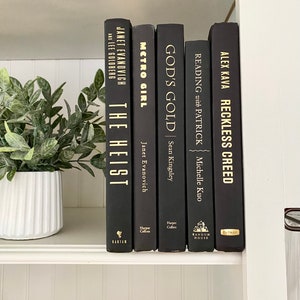 May include: A collection of five hardcover books with black covers and gold lettering. Titles include "The Heist," "Metro Girl," "God's Gold," "Reading with Patrick," and "Reckless Creed." The books are arranged on a white shelf with a potted plant.