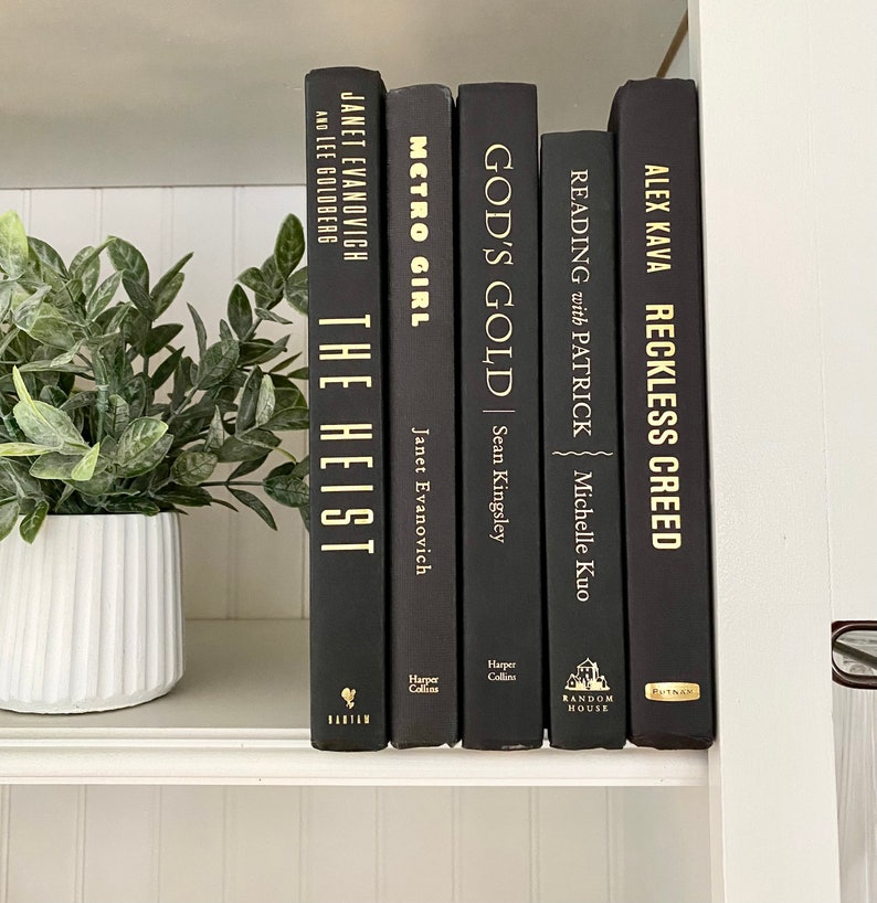 May include: A collection of five hardcover books with black covers and gold lettering. Titles include "The Heist," "Metro Girl," "God's Gold," "Reading with Patrick," and "Reckless Creed." The books are arranged on a white shelf with a potted plant.