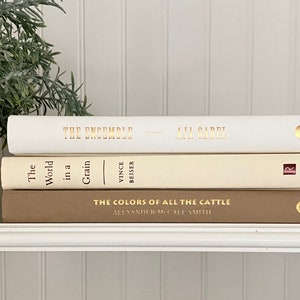 May include: A stack of three books with gold lettering on the spines. The top book is white with the title "The Ensemble" and the author "Ana Garri". The middle book is cream with the title "The World in a Grain" and the author "Vince Beiser". The bottom book is brown with the title "The Colors of All the Cattle" and the author "Alexander McCall Smith".