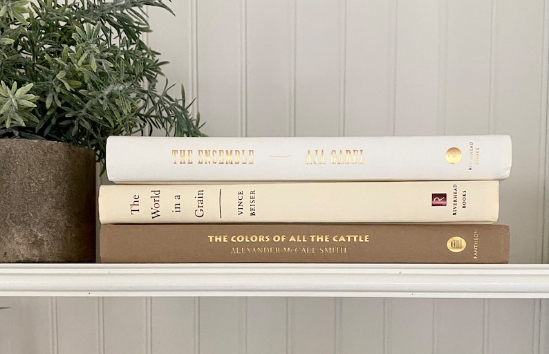 May include: A stack of three books with gold lettering on the spines. The top book is white with the title "The Ensemble" and the author "Ana Garri". The middle book is cream with the title "The World in a Grain" and the author "Vince Beiser". The bottom book is brown with the title "The Colors of All the Cattle" and the author "Alexander McCall Smith".