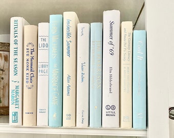 May include: A collection of hardcover books with various titles and authors, arranged on a white shelf. The books feature a palette of soft colors, including white, light blue, and cream, creating a cohesive and aesthetically pleasing display. The spines display the titles.
