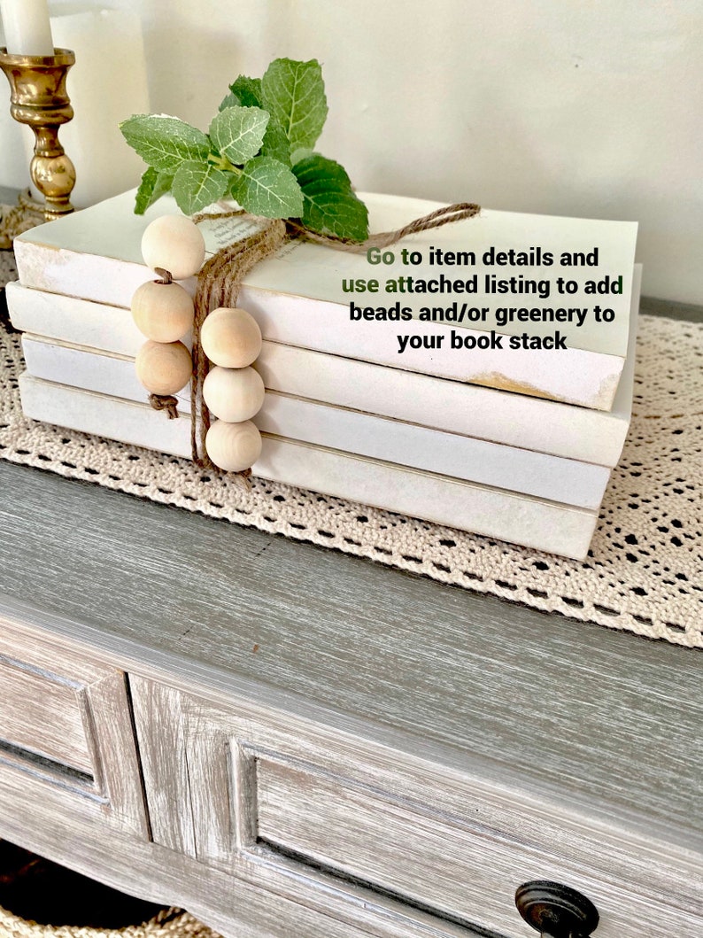 May include: A stack of white books with a brown twine bow and wooden beads. A sprig of green foliage is tucked into the twine. The text "Go to item details and use attached listing to add beads and/or greenery to your book stack" is written on a white background.