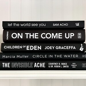 May include: A stack of five black hardcover books with white lettering. Titles include "let the world see you", "ON THE COME UP", "CHILDREN OF EDEN", "CIRCLE IN THE WATER", and "THE INVISIBLE ACHE". The books are arranged on a white shelf.