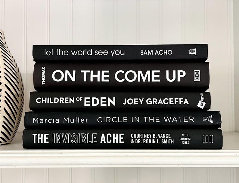 May include: A stack of five black hardcover books with white lettering. Titles include "let the world see you", "ON THE COME UP", "CHILDREN OF EDEN", "CIRCLE IN THE WATER", and "THE INVISIBLE ACHE". The books are arranged on a white shelf.