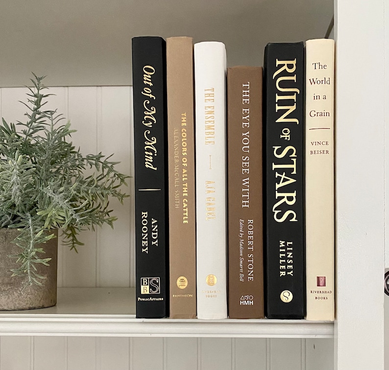 May include: A bookshelf with a variety of books, including "Out of My Mind" by Andy Rooney, "The Colors of All the Cattle" by Alexander McCall Smith, "The Ensemble" by Ava Wanel, "The Eye You See With" by Robert Stone, "Ruin of Stars" by Linsey Miller, and "The World in a Grain" by Vince Beiser.