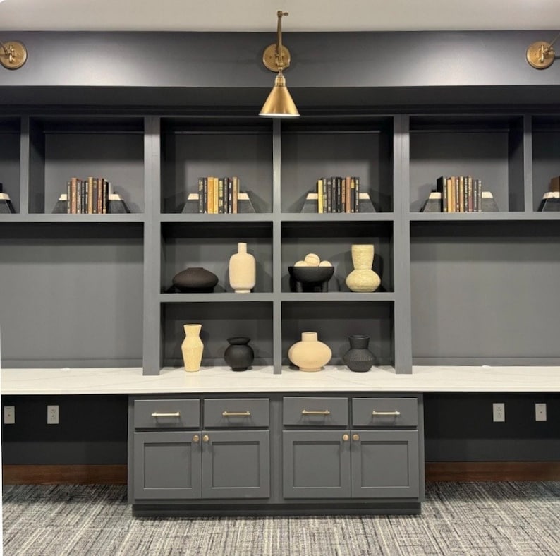 May include: A dark gray built-in bookshelf with white countertops and gray cabinets. The shelves are filled with books and decorative vases. The room has a modern design.