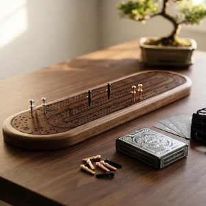 Könnte beinhalten: Ein ovales Cribbage-Spielbrett aus Holz mit einer dunklen, vertieften Bahn und nummerierten Löchern. Das Set enthält zwei silberne, zwei schwarze und zwei kupferfarbene Metallstifte im Brett sowie weitere lose Stifte, ein Kartenspiel und einen schwarzen Beutel.