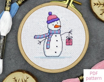 May include: A cross stitch pattern of a snowman holding a gift box. The snowman is wearing a pink and purple hat and a blue and pink striped scarf. The pattern is in a wooden embroidery hoop. The text "SNOWMAN 3" is at the top of the image. The text "PDF pattern" is at the bottom of the image.