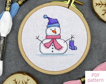 May include: A cross-stitch pattern of a snowman wearing a purple hat and pink scarf. The snowman is holding a blue sock. The pattern is in a wooden embroidery hoop. The text "SNOWMAN 2" is at the top of the image. The text "PDF pattern" is at the bottom of the image.