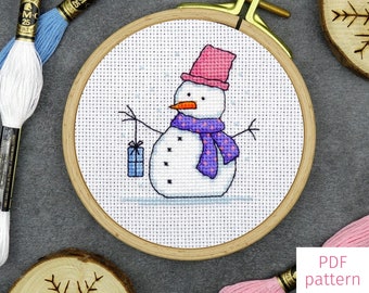 May include: A cross-stitch pattern of a snowman holding a gift box. The snowman is wearing a pink hat and a purple scarf with white polka dots. The pattern is displayed in a wooden embroidery hoop.