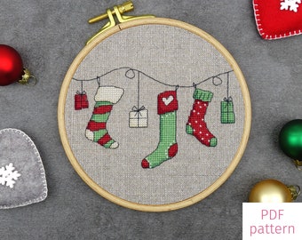 May include: A cross-stitch pattern for hanging stockings. The pattern shows three stockings hanging from a line with presents and hearts. The pattern is in a wooden embroidery hoop.