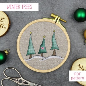 Puede incluir: Un bastidor de bordado de punto de cruz con tres árboles de Navidad verdes, cada uno coronado con una estrella y un corazón rosa. El texto "WINTER TREES" está en la parte superior y "PDF pattern" en la inferior. Decoraciones navideñas y rodajas de madera rodean el bastidor.