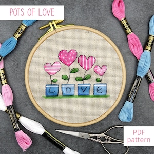 Puede incluir: Un patrón de punto de cruz con cinco corazones rosas en macetas que deletrean la palabra "LOVE". El patrón se muestra en un aro de bordado de madera con carretes de hilo de bordar de colores que lo rodean.