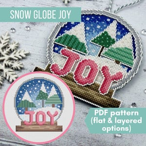 Op de afbeelding: Een kruissteek kerstornament met een sneeuwbolontwerp. Het ornament toont een blauwe lucht, groene bomen en het woord "JOY" in roze. De tekst "SNOW GLOBE JOY" staat bovenaan. Een kleinere versie van het ontwerp staat linksonder.