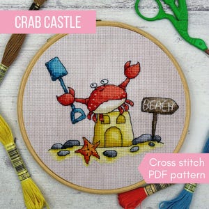 May include: A cross-stitch embroidery hoop featuring a crab holding a blue shovel, standing on a yellow sandcastle. The design includes a starfish, rocks, and a sign that says "Beach". The text "Crab Castle" is at the top.