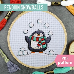 May include: Cross-stitch embroidery hoop featuring a cartoon penguin with a red, green, and white scarf, juggling snowballs. The text "PENGUIN SNOWBALLS" is at the top, and "PDF pattern" is in a pink banner. Embroidery floss surrounds the hoop.
