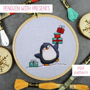 May include: A cross-stitch pattern featuring a penguin holding a stack of presents. The pattern is displayed in a wooden embroidery hoop with a light gray background. The penguin is black and white with a red beak and orange feet. The presents are wrapped in red, green, and blue paper with white polka dots. The text "PDF pattern" is displayed in the lower right corner of the image.