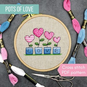 May include: A round wooden embroidery hoop containing a cross-stitch design of four heart-shaped flowers in blue pots spelling the word LOVE. The design is on beige fabric, surrounded by colorful embroidery floss skeins and metal scissors.