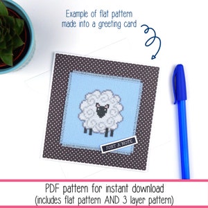 Cross stitch of a small stylised sheep stitched on blue Aida and finished as a greeting card with a potted plant and pen peeking in at the sides of the photo. The listing is for the PDF cross stitch pattern.