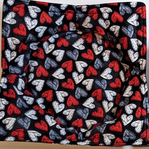 May include: Two square fabric bowl cozies with a black background and a pattern of red, white, and gray hearts. The cozies are made of a soft, quilted fabric.