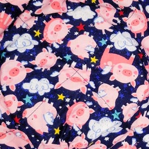 May include: Two square fabric bowl cozies with a navy blue background. The fabric is patterned with pink pigs, white clouds, yellow stars, and red accents. The cozies are quilted and have a folded design.