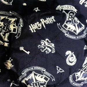 May include: Two navy blue, square bowl cozies with a quilted pattern. The fabric is printed with white Harry Potter themed designs, including the Hogwarts crest, stars, and text. These cozies are designed to hold bowls.