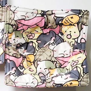 May include: A set of four quilted pot holders with a repeating pattern of cartoon cats in various colors. The pot holders are square-shaped with a gray border and a quilted design.