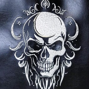 May include: Black tote bag with a white embroidered skull design. The skull has detailed features and is surrounded by ornate, swirling embellishments. The bag is made of a textured material.