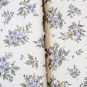 May include: A cream-colored floral print wallet and matching wristlet. The fabric features a pattern of lavender and green flowers. The wallet is folded, and the wristlet has a silver-colored key ring.